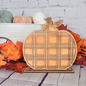 May include: A wooden pumpkin cutout with a plaid pattern. The pumpkin is standing on a small wooden base. The pumpkin is orange and the base is brown. The pumpkin is surrounded by fall leaves.