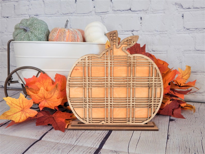 May include: A wooden pumpkin cutout with a plaid pattern. The pumpkin is standing on a small wooden base. The pumpkin is orange and the base is brown. The pumpkin is surrounded by fall leaves.