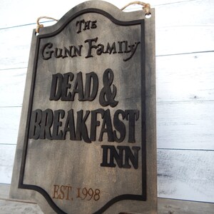 May include: A wooden sign with a distressed finish, featuring the text "The Gunn Family Dead & Breakfast Inn Est. 1998". The sign has a black border and a rope for hanging.