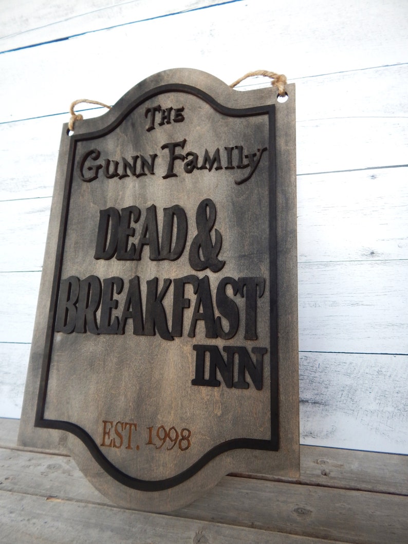 May include: A wooden sign with a distressed finish, featuring the text "The Gunn Family Dead & Breakfast Inn Est. 1998". The sign has a black border and a rope for hanging.