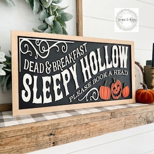 May include: A rectangular wooden sign with a light wood frame and a black background. The sign features white 3D lettering that reads DEAD & BREAKFAST SLEEPY HOLLOW PLEASE BOOK A HEAD, accompanied by three orange pumpkins and white scrollwork.