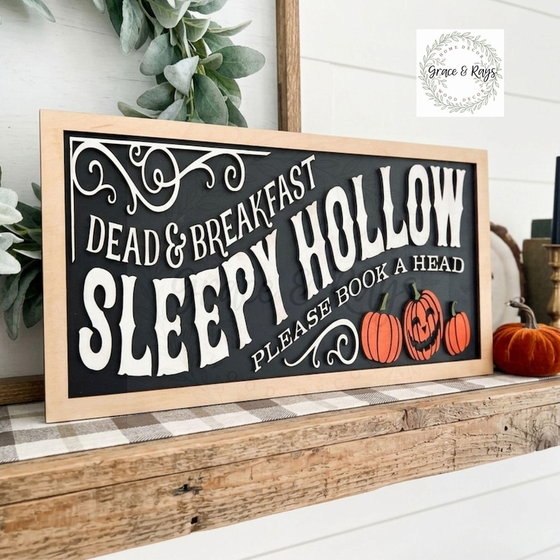 May include: A rectangular wooden sign with a light wood frame and a black background. The sign features white 3D lettering that reads DEAD & BREAKFAST SLEEPY HOLLOW PLEASE BOOK A HEAD, accompanied by three orange pumpkins and white scrollwork.