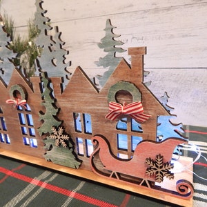 May include: Wooden Christmas village scene with houses, trees, and a sleigh with lights. The houses are painted in a rustic brown with white accents. The trees are green with white snow. The sleigh is red with white accents.