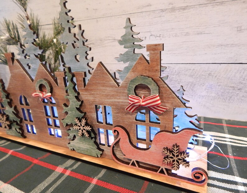 May include: Wooden Christmas village scene with houses, trees, and a sleigh with lights. The houses are painted in a rustic brown with white accents. The trees are green with white snow. The sleigh is red with white accents.