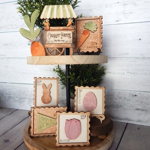 May include: A tiered wooden stand with a green plant and a wooden carrot patch sign on the top tier. The bottom tier has four wooden stamps with Easter and spring themes. The stamps feature a bunny, an egg, a tulip, and a banner that says "Spring".