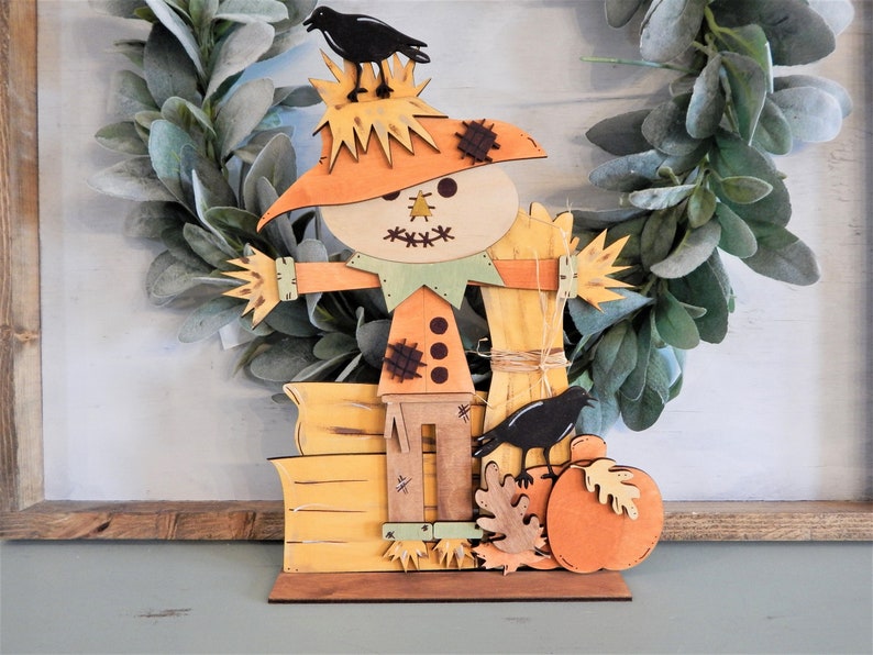 May include: Wooden fall-themed decoration featuring a scarecrow with a straw hat and crow perched on top. The scarecrow has an orange body, green collar, and wooden accents. A pumpkin, leaves, and another crow are also included in the design. The decoration is approximately 10 inches tall.