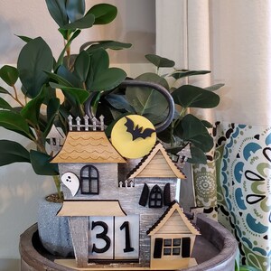 Puede incluir: Decoraci&oacute;n de casa de Halloween de madera con una combinaci&oacute;n de colores gris y beige. La casa presenta un murci&eacute;lago y una luna, un fantasma y un calendario que muestra el n&uacute;mero 31. La decoraci&oacute;n est&aacute; colocada en una bandeja de madera.