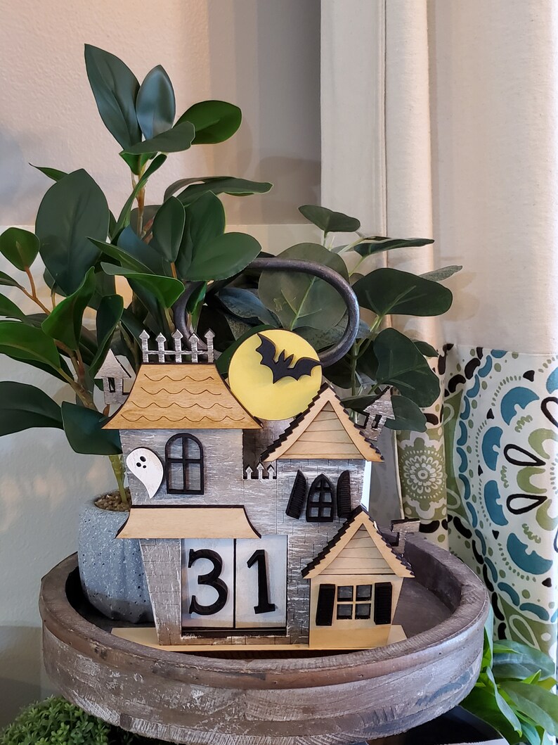 Puede incluir: Decoraci&oacute;n de casa de Halloween de madera con una combinaci&oacute;n de colores gris y beige. La casa presenta un murci&eacute;lago y una luna, un fantasma y un calendario que muestra el n&uacute;mero 31. La decoraci&oacute;n est&aacute; colocada en una bandeja de madera.