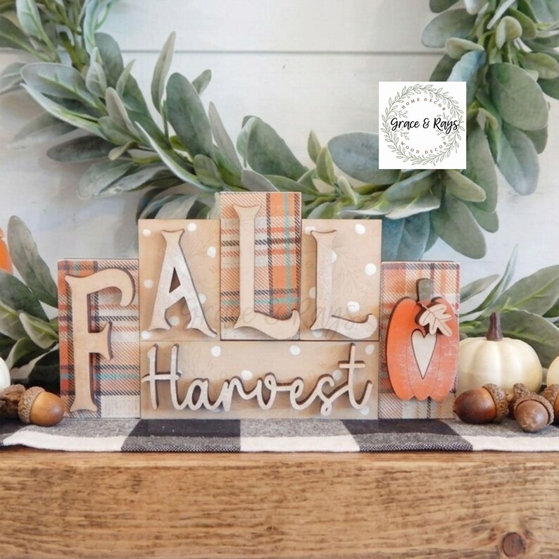 May include: A set of wooden blocks spelling FALL HARVEST with a plaid pattern, white polka dots, and a cutout pumpkin shape. The blocks feature light wood lettering and a small leaf accent. A logo for Grace & Rays is visible in the upper right corner.