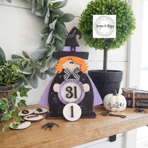 K&ouml;nnte beinhalten: Ein h&ouml;lzernes Halloween-Countdown-Schild in Form einer Hexe mit orangefarbenen Haaren, einem schwarzen Hut und einem violetten Umhang. Das Schild hat eine kreisf&ouml;rmige Mitte mit der Zahl 31 und dem Text Countdown to Halloween sowie eine separate runde Holzzahl 1.