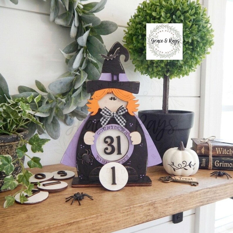 K&ouml;nnte beinhalten: Ein h&ouml;lzernes Halloween-Countdown-Schild in Form einer Hexe mit orangefarbenen Haaren, einem schwarzen Hut und einem violetten Umhang. Das Schild hat eine kreisf&ouml;rmige Mitte mit der Zahl 31 und dem Text Countdown to Halloween sowie eine separate runde Holzzahl 1.