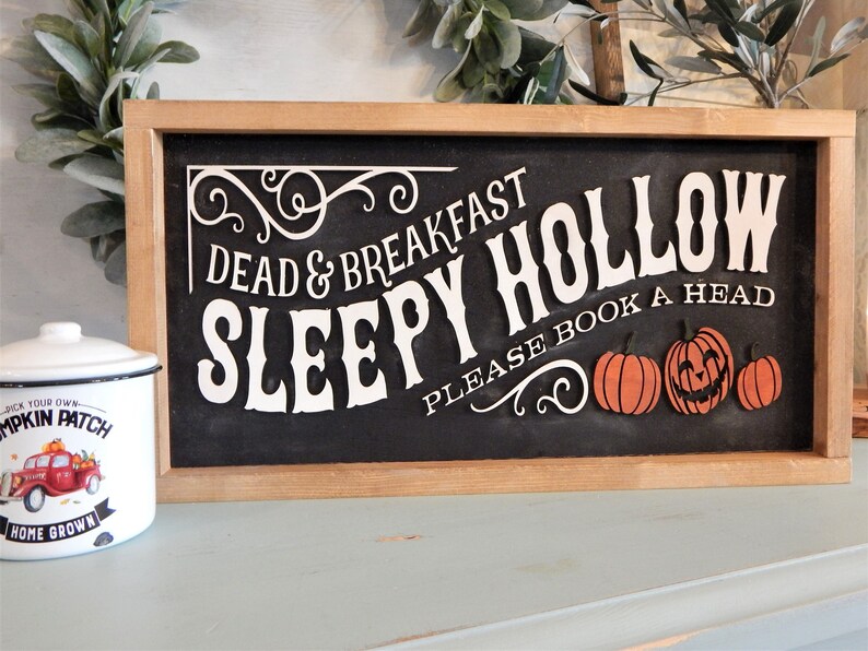 May include: A rectangular wooden sign with a black background and white text reading Dead & Breakfast Sleepy Hollow Please Book A Head, featuring three orange pumpkins, one with a carved face, and a white enamel canister with a red truck graphic.