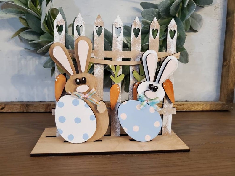 Pu&ograve; includere: Decorazione pasquale in legno con due figure di coniglietti che tengono uova a pois, ambientata davanti a una staccionata bianca con cuori intagliati. Sono incluse anche carote e una ghirlanda. La base &egrave; in legno marrone chiaro.