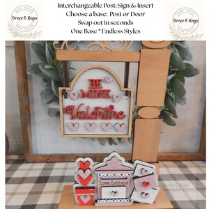 May include: A wooden Valentine's Day sign with the words "Be Mine Valentine" in red and pink. The sign has a decorative frame and heart accents. The base includes a mailbox that says "Love Letters".