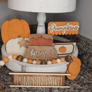 May include: A white tiered tray decorated with autumn-themed wooden cutouts. The tray features a pumpkin, a truck with the word "Grateful" on the side, a pumpkin stand with the word "Pumpkins" on top, and a sign that reads "Pumpkin Patch". The tray is also decorated with a beaded garland with orange, white, and brown beads.