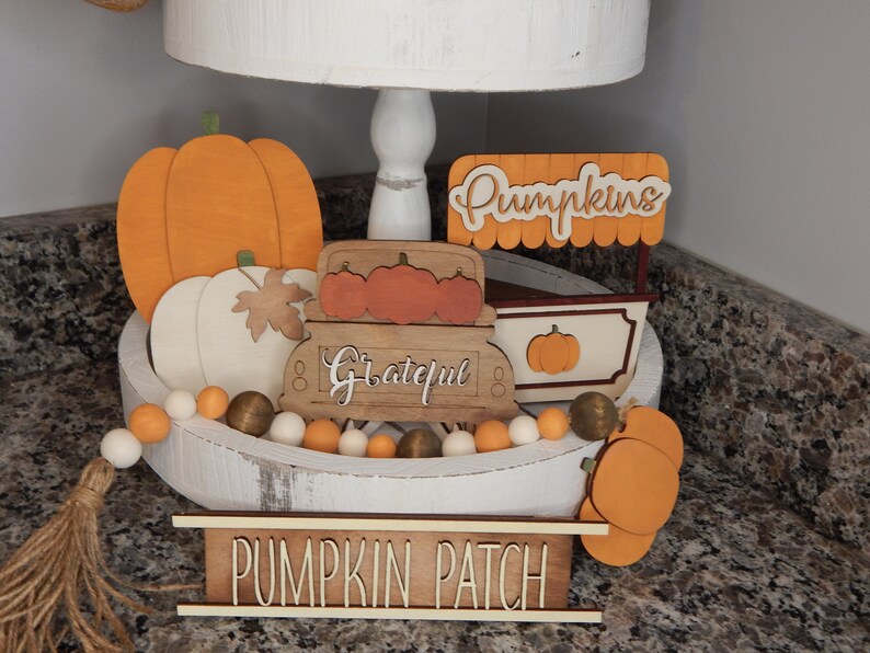 May include: A white tiered tray decorated with autumn-themed wooden cutouts. The tray features a pumpkin, a truck with the word "Grateful" on the side, a pumpkin stand with the word "Pumpkins" on top, and a sign that reads "Pumpkin Patch". The tray is also decorated with a beaded garland with orange, white, and brown beads.
