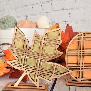 May include: Two wooden fall decorations, a pumpkin and a maple leaf, with a plaid pattern. The decorations are on a wooden stand and are painted in orange, green, and brown.