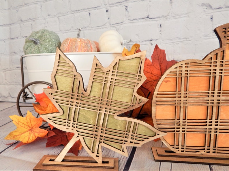 May include: Two wooden fall decorations, a pumpkin and a maple leaf, with a plaid pattern. The decorations are on a wooden stand and are painted in orange, green, and brown.