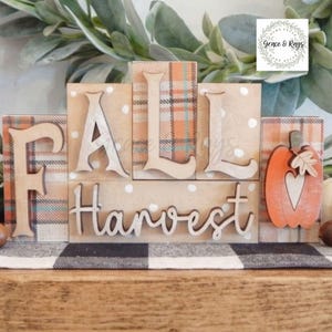 May include: A wooden block sign featuring the text FALL HARVEST with a pumpkin cutout. The blocks have a tan background with white polka dots and orange, blue, and white plaid patterns. The letters are raised and light-colored wood.