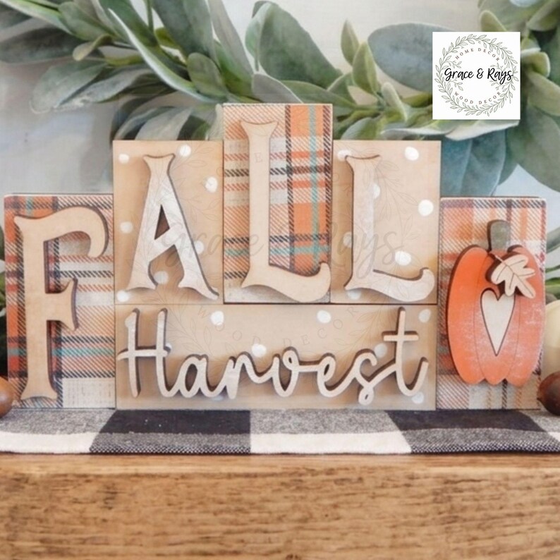 May include: A wooden block sign featuring the text FALL HARVEST with a pumpkin cutout. The blocks have a tan background with white polka dots and orange, blue, and white plaid patterns. The letters are raised and light-colored wood.