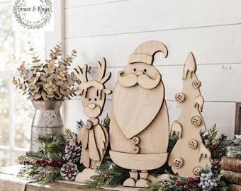 May include: Three light-colored wooden holiday decorations featuring a reindeer, a Santa Claus figure, and a Christmas tree adorned with small wooden buttons. The figures are arranged together on a surface with greenery and pinecones.