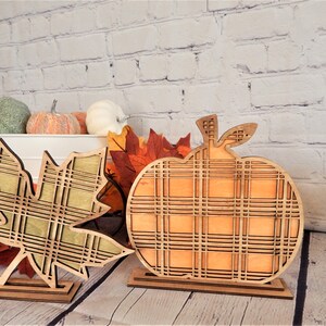 May include: Two wooden fall decorations, a pumpkin and a leaf, both with a plaid pattern. The pumpkin is orange and the leaf is green. Both are standing on small wooden bases.