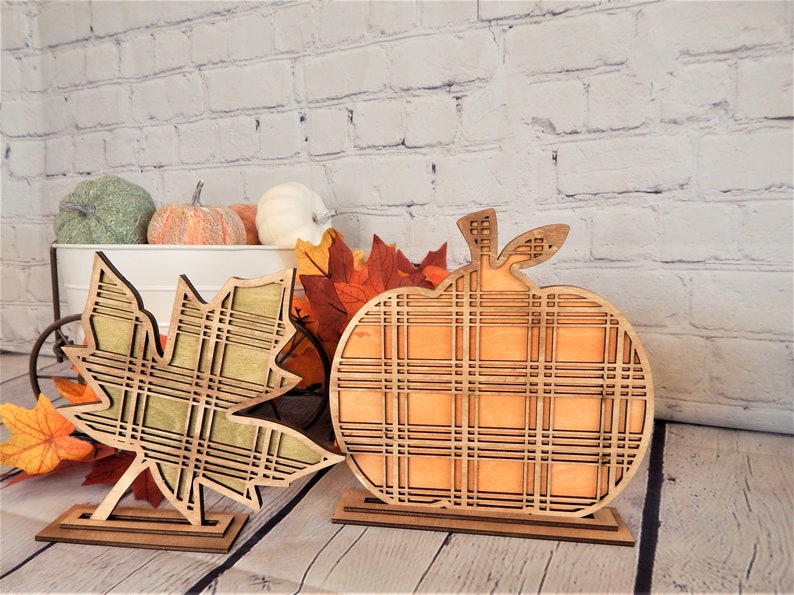 May include: Two wooden fall decorations, a pumpkin and a leaf, both with a plaid pattern. The pumpkin is orange and the leaf is green. Both are standing on small wooden bases.