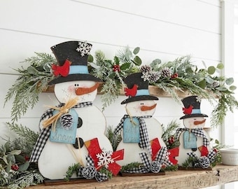 May include: Three white wooden snowman figures of varying sizes, each wearing a black top hat with a light blue band, a red cardinal, and a white snowflake. Each snowman features a carrot nose, black buttons, and a blue tag reading Let it Snow.