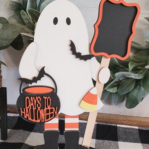 May include: A wooden ghost holding a chalkboard sign and a cauldron with the text "Days to Halloween". The ghost is wearing black boots and orange and white striped stockings. The ghost is standing on a black base.