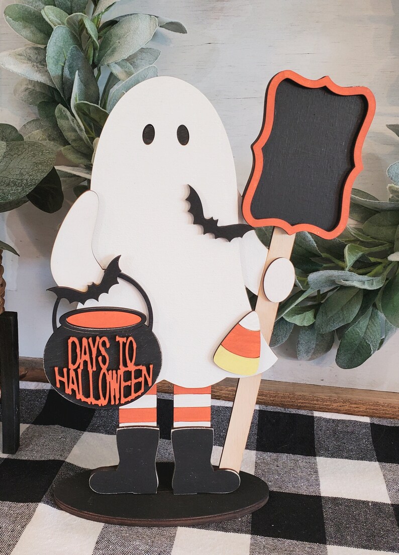 May include: A wooden ghost holding a chalkboard sign and a cauldron with the text "Days to Halloween". The ghost is wearing black boots and orange and white striped stockings. The ghost is standing on a black base.