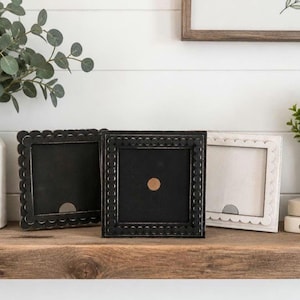 May include: Three square picture frames with decorative edges sit on a wooden shelf. Two are black, and one is white. A small plant in a metal pot and a white vase with greenery are also on the shelf. The frames are empty.