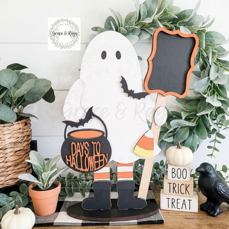 May include: A white wooden ghost cutout standing on a base, holding a black cauldron sign that reads DAYS TO HALLOWEEN and a black chalkboard sign. The ghost wears orange and white striped leggings with black boots and holds a candy corn cutout.