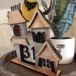 Puede incluir: Calendario de Halloween de madera con forma de casa embrujada. La casa tiene una combinaci&oacute;n de colores gris y marr&oacute;n, con una luna llena y un recorte de murci&eacute;lago. El calendario muestra el n&uacute;mero 31. La casa est&aacute; sobre una bandeja de madera.