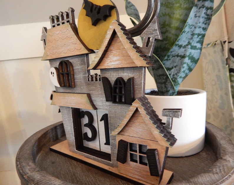 Puede incluir: Calendario de Halloween de madera con forma de casa embrujada. La casa tiene una combinaci&oacute;n de colores gris y marr&oacute;n, con una luna llena y un recorte de murci&eacute;lago. El calendario muestra el n&uacute;mero 31. La casa est&aacute; sobre una bandeja de madera.