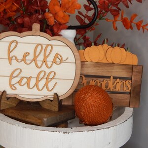 May include: A white wooden pumpkin sign with the words "hello fall" in orange lettering, a small wooden crate with the word "pumpkins" in orange lettering, and a small orange woven pumpkin on a white tiered tray.