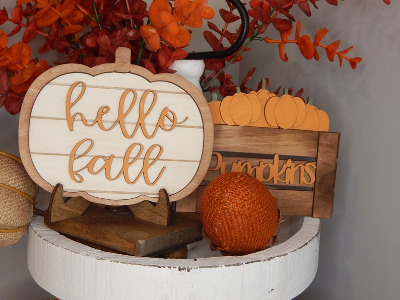 May include: A white wooden pumpkin sign with the words "hello fall" in orange lettering, a small wooden crate with the word "pumpkins" in orange lettering, and a small orange woven pumpkin on a white tiered tray.