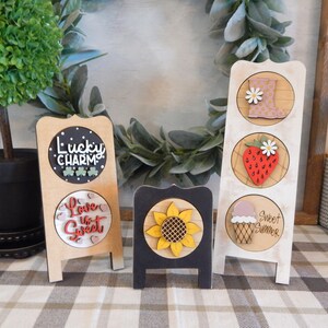 May include: Three wooden signs with different designs. The first sign has a black circle with white polka dots and the text "Lucky Charm" in red and green. The second sign has a red circle with white hearts and the text "Love is Sweet" in red. The third sign has three circles with a pink rain boot, a red strawberry, and a pink ice cream cone. The text "Sweet Summer" is written on the sign.