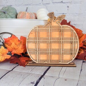 May include: A wooden pumpkin cutout with a plaid pattern. The pumpkin is orange and has a brown stem. It is sitting on a wooden stand.