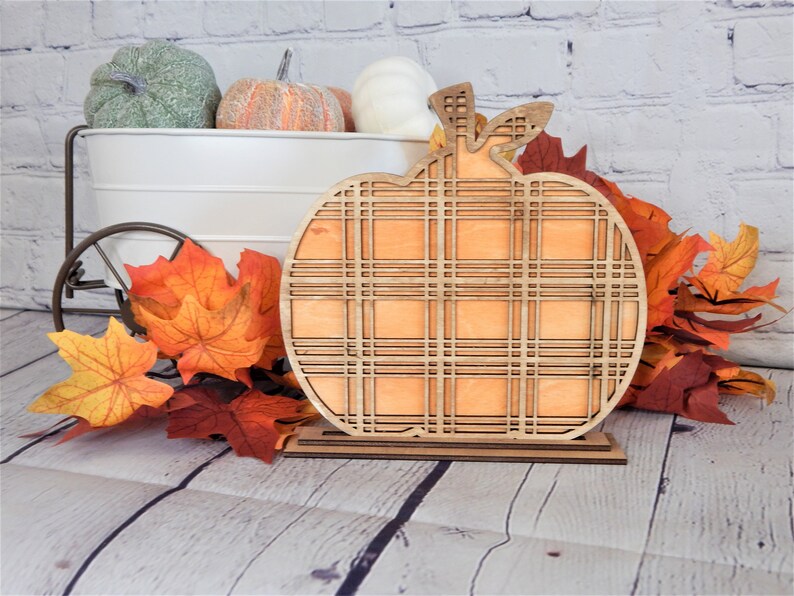 May include: A wooden pumpkin cutout with a plaid pattern. The pumpkin is orange and has a brown stem. It is sitting on a wooden stand.