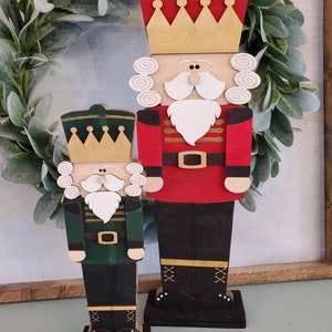 May include: Two wooden nutcracker figurines, one red and one green, with gold crowns and white beards. They are standing on black bases with gold accents.