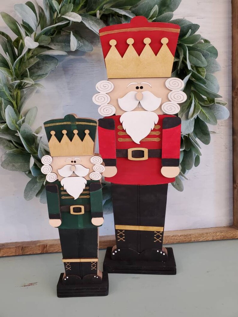 May include: Two wooden nutcracker figurines, one red and one green, with gold crowns and white beards. They are standing on black bases with gold accents.