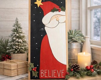 May include: Wooden Christmas sign featuring a partial Santa Claus design with a red hat and coat, white beard, and a yellow star. The word "BELIEVE" is written in white letters. The background is black with white dots.
