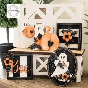 May include: A collection of Halloween-themed wooden decor items including two white ghost figures holding orange pumpkins, a square sign with black and white stripes and an orange flower, a square sign with the word Boo, and an oval sign with a ghost.