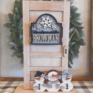 Puede incluir: Decoraci&oacute;n de puerta de madera con tem&aacute;tica de mu&ntilde;eco de nieve. La puerta es de color marr&oacute;n claro con un ojo de cerradura y un letrero que dice "In the making we are building a snowman". Tres mu&ntilde;ecos de nieve est&aacute;n en la base de la puerta.