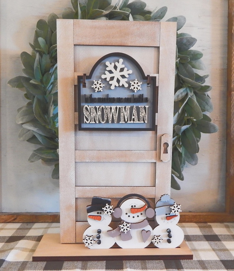 Puede incluir: Decoraci&oacute;n de puerta de madera con tem&aacute;tica de mu&ntilde;eco de nieve. La puerta es de color marr&oacute;n claro con un ojo de cerradura y un letrero que dice "In the making we are building a snowman". Tres mu&ntilde;ecos de nieve est&aacute;n en la base de la puerta.