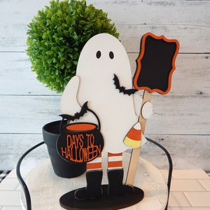 May include: White ghost figurine holding a black cauldron with the text "Days to Halloween" and a black chalkboard sign. The ghost is wearing red and white striped stockings and black boots. The figurine is standing on a wooden base.