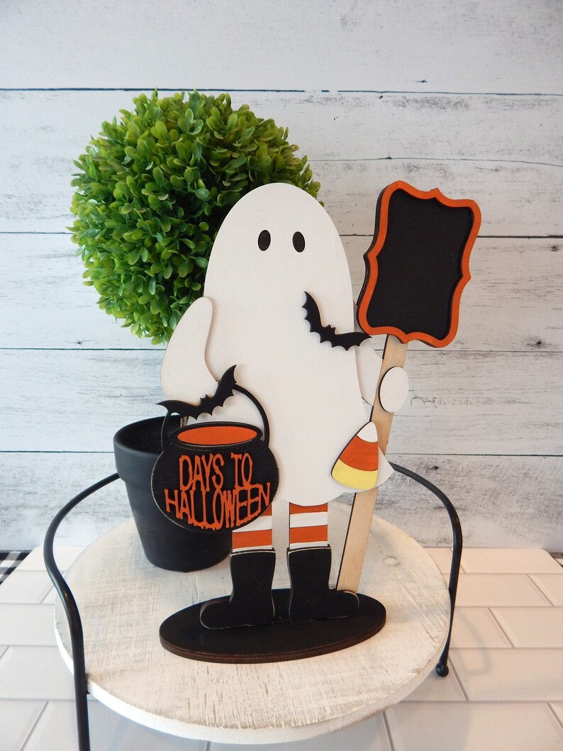 May include: White ghost figurine holding a black cauldron with the text "Days to Halloween" and a black chalkboard sign. The ghost is wearing red and white striped stockings and black boots. The figurine is standing on a wooden base.