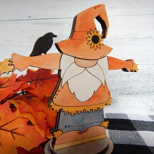 May include: A wooden gnome figurine dressed as a scarecrow with a straw hat, orange shirt, blue jeans, and a white beard. The gnome is holding out its arms and has a black crow perched on its arm. The gnome is standing on a wooden base and is surrounded by fall leaves.