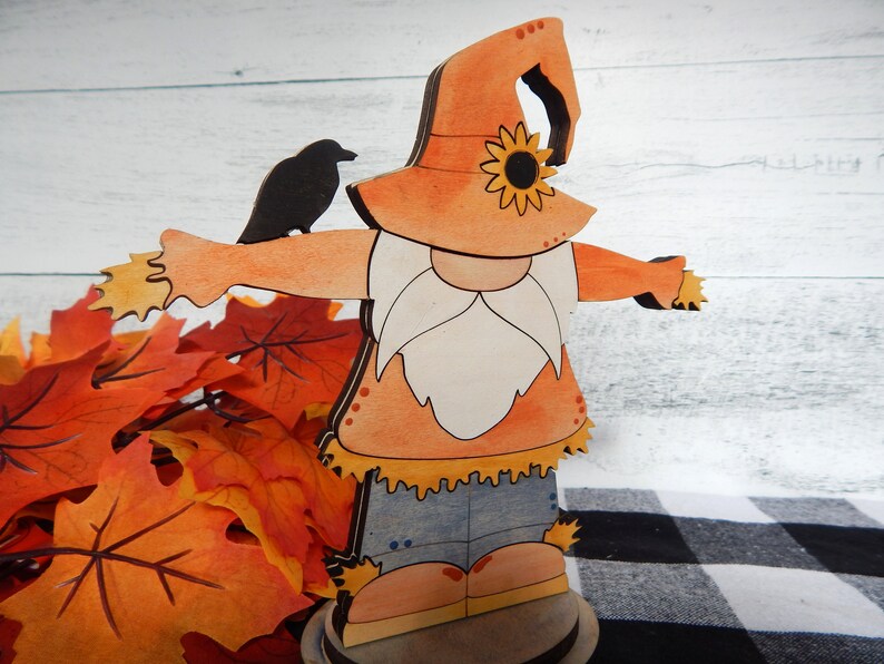 May include: A wooden gnome figurine dressed as a scarecrow with a straw hat, orange shirt, blue jeans, and a white beard. The gnome is holding out its arms and has a black crow perched on its arm. The gnome is standing on a wooden base and is surrounded by fall leaves.