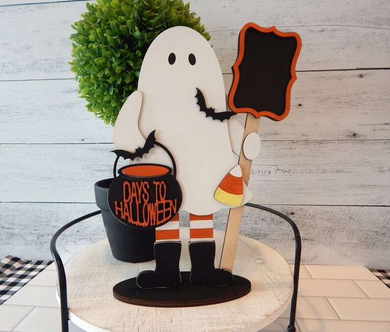 May include: A white ghost holding a black cauldron with the text "Days to Halloween" and a chalkboard sign. The ghost is wearing black boots and red and white striped legs. The ghost is standing on a wooden base.